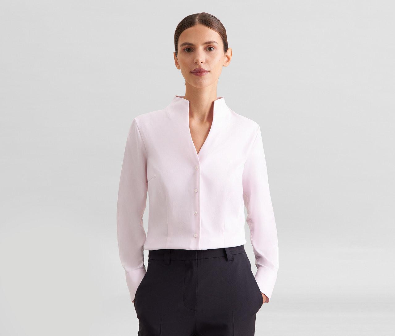 Femme portant une chemise rose pâle à manches longues avec col montant et un pantalon noir, regardant la caméra.