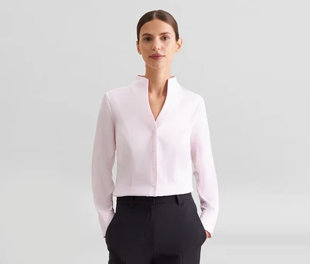 Femme portant une chemise rose pâle à manches longues avec col montant et un pantalon noir, regardant la caméra.