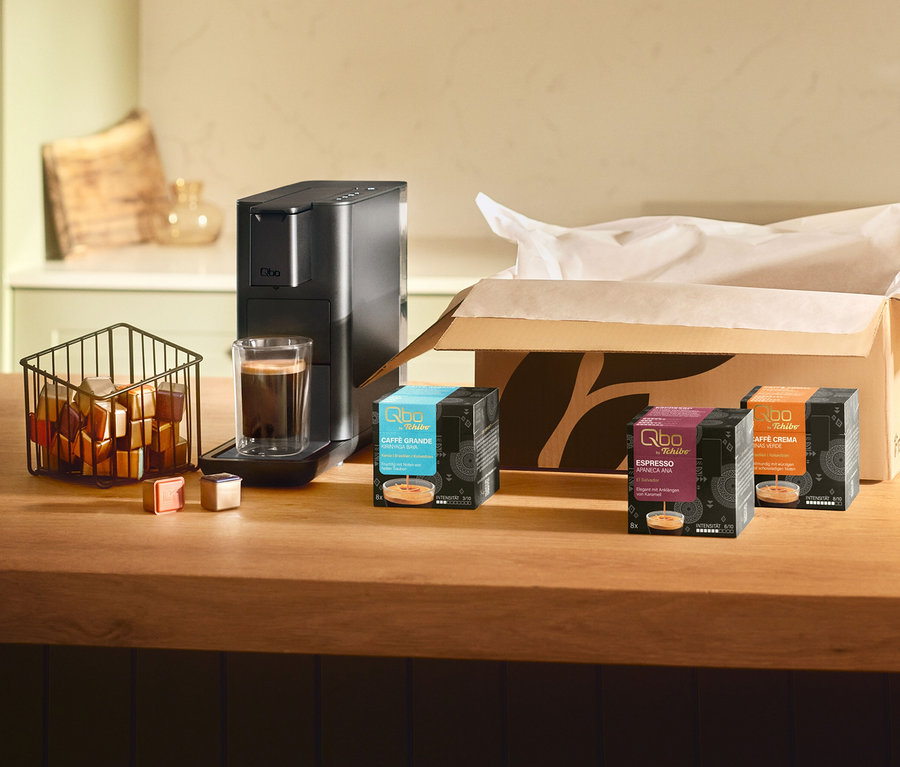 Machine à café Qbo Iconic Dark Chrome noire avec café dans un verre, un panier de capsules et trois boîtes de capsules Qbo: Qbo Caffè Grande KIRINYAGA BAYA, une boîte Qbo Espresso violette et Qbo Caffè Minas Verde.