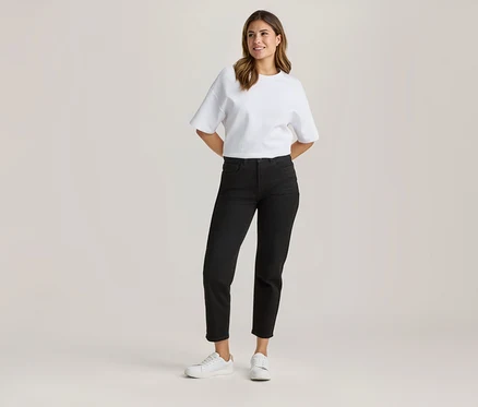 Femme debout, portant un t-shirt blanc ample, un jean noir raccourci et des baskets blanches, les mains derrière le dos et souriante.