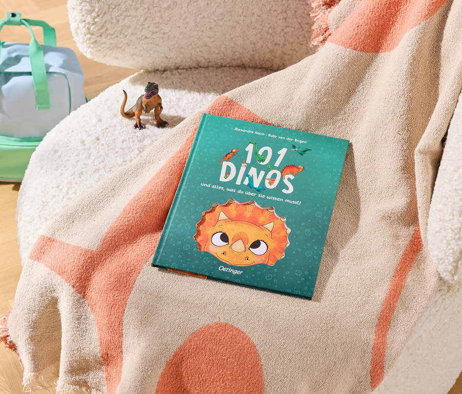 Un sac à dos bleu clair et vert menthe est posé sur un parquet en bois à côté d'un fauteuil crème. Une couverture beige et orange est drapée sur le fauteuil, avec un petit jouet dinosaure marron et un livre turquoise intitulé « 101 DINOS ».