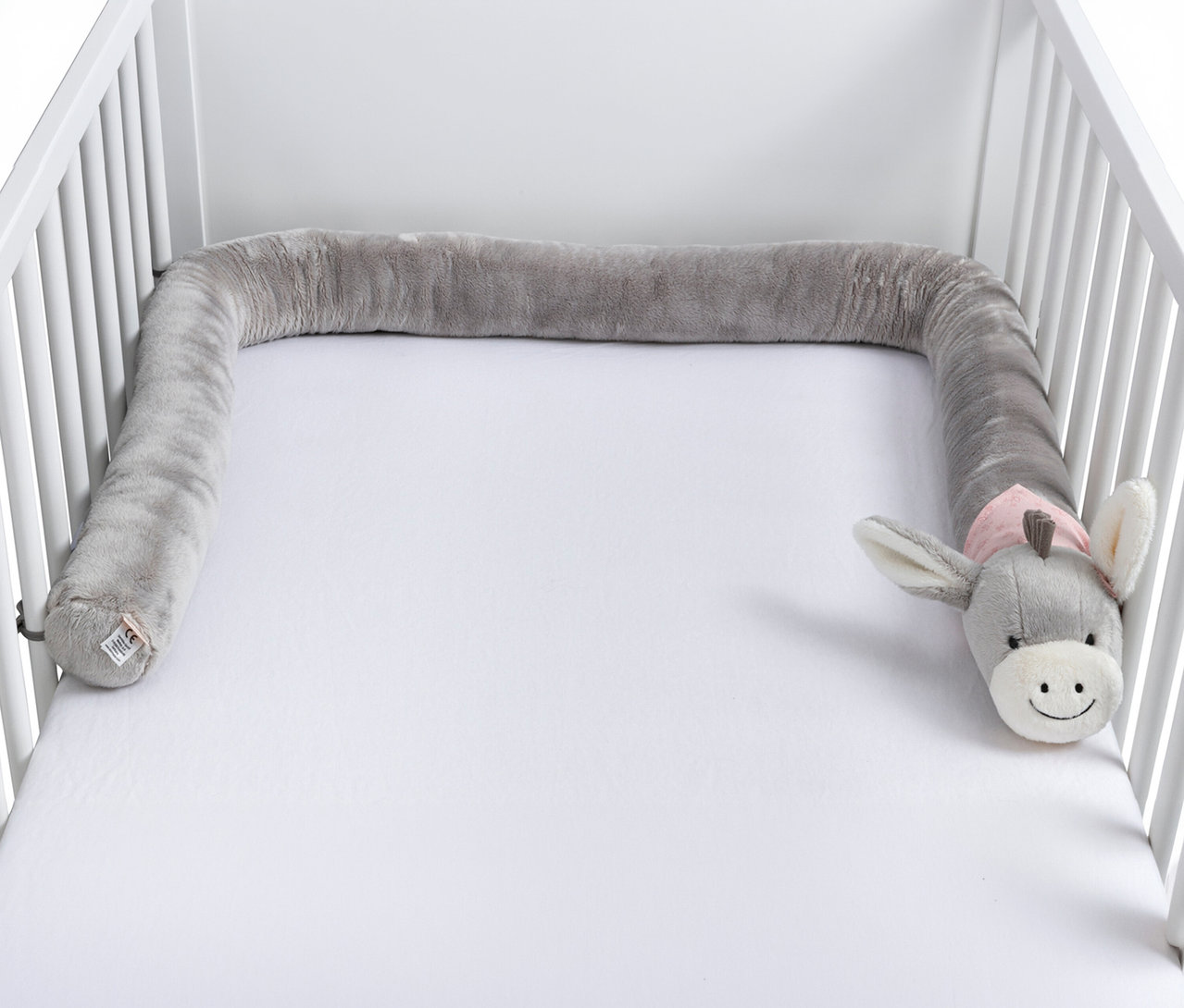 Bumper de lit gris en peluche en forme de serpent avec une tête d'âne, posé dans un lit de bébé blanc.