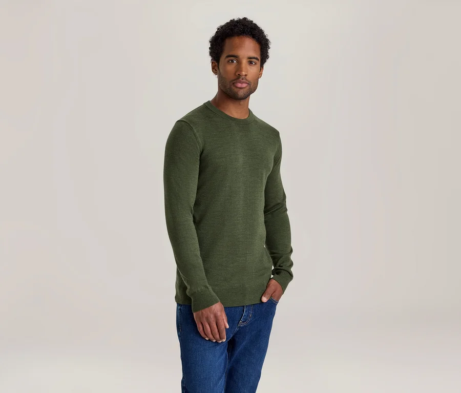 Homme aux cheveux bouclés foncés portant un t-shirt vert à manches longues et un jean bleu, une main dans la poche.