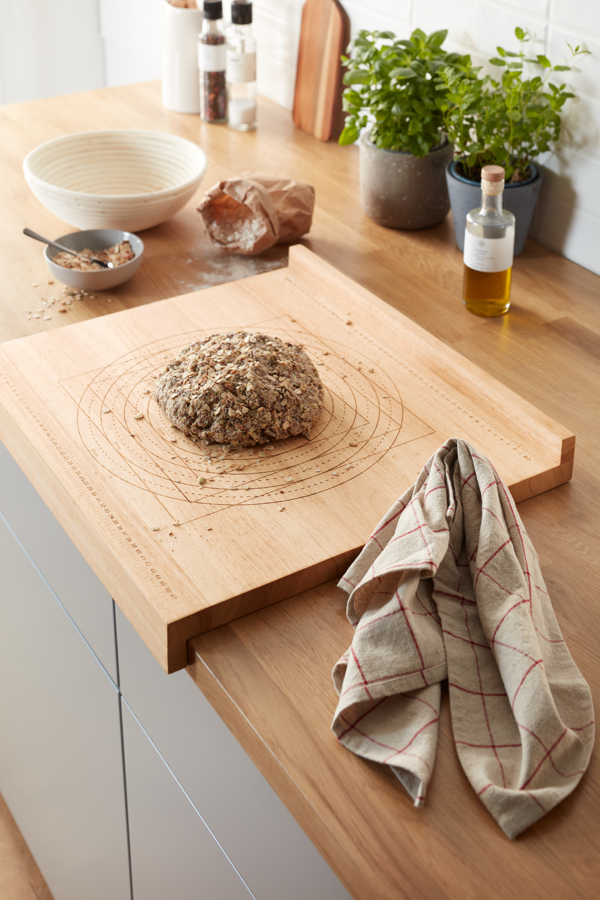 Sur un comptoir de cuisine, une planche à découper et plan de travail en bois avec une miche de pain. Des herbes en pot et d'autres ustensiles de cuisine sont visibles en arrière-plan.