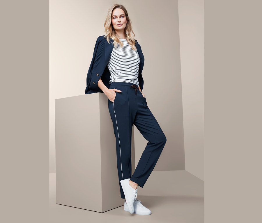 Femme blonde posant en blazer bleu marine et pantalon de jogging.