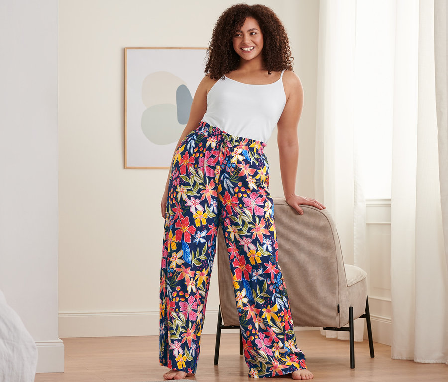 Une femme appuyée sur une chaise porte un caraco blanc et un pantalon relax bleu foncé à motifs floraux.