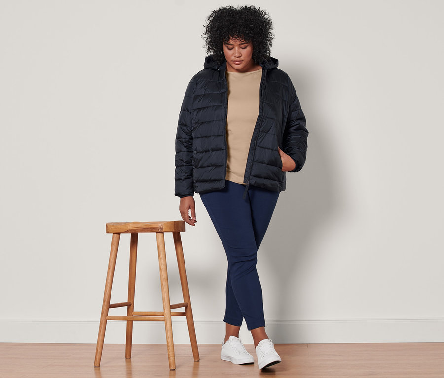 Une femme debout porte une veste matelassée à capuche bleu foncé, un t-shirt manches 3/4 beige et un pantalon stretch bleu foncé.