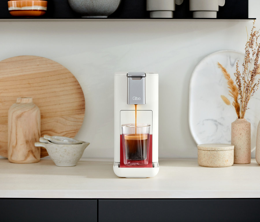 Une machine à café Qbo ESSENTIAL Bright Coral verse du café dans un verre. Des objets décoratifs sont visibles en arrière-plan et une Qbo ICONIC Dark Chrome.