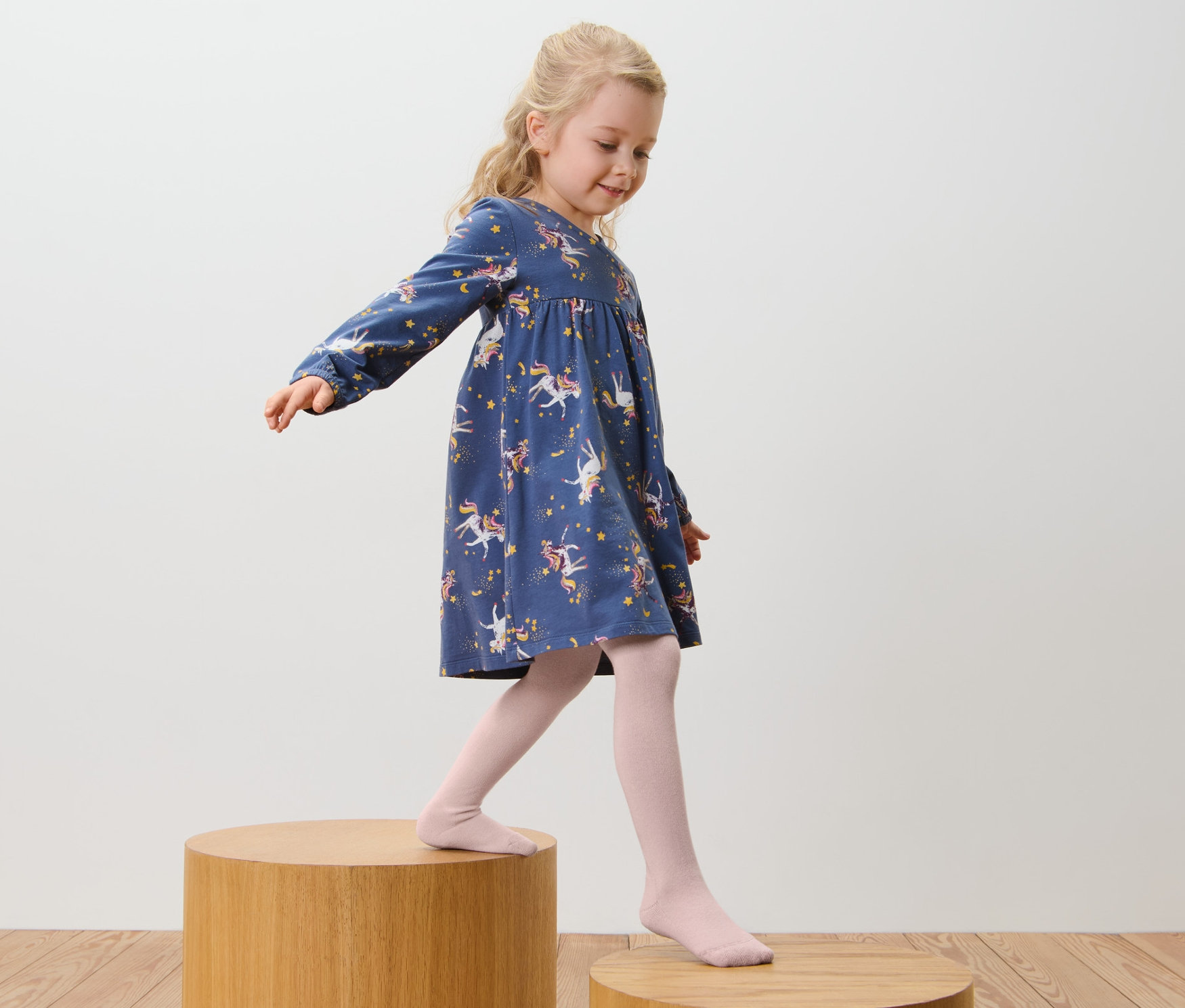Une fille portant une robe bleue à imprimé licorne et des collants en coton roses, lot de 5, rose et marron, marche sur des marches en bois.