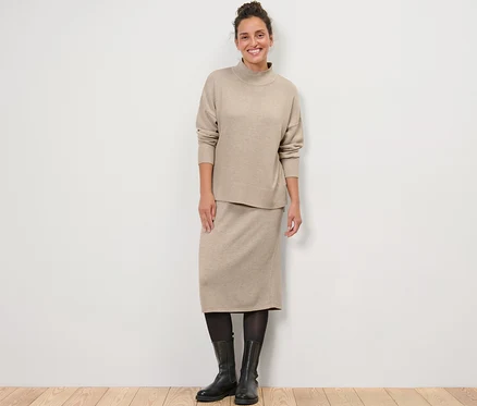 Une femme debout portant un pull en maille fine avec col montant et une jupe tricotée.