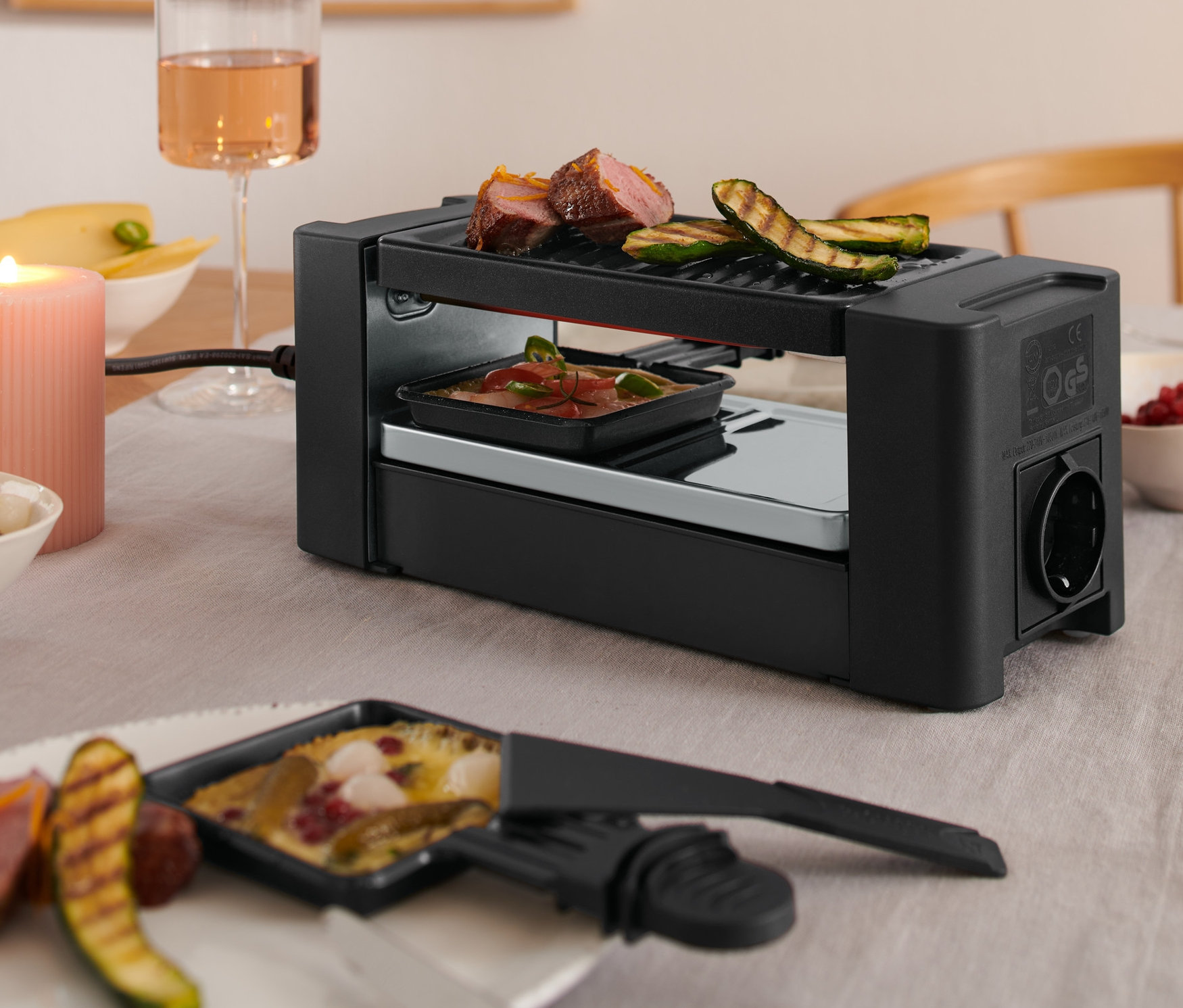 Gros plan d'une table avec un four à raclette pour deux, un verre de vin et des aliments.