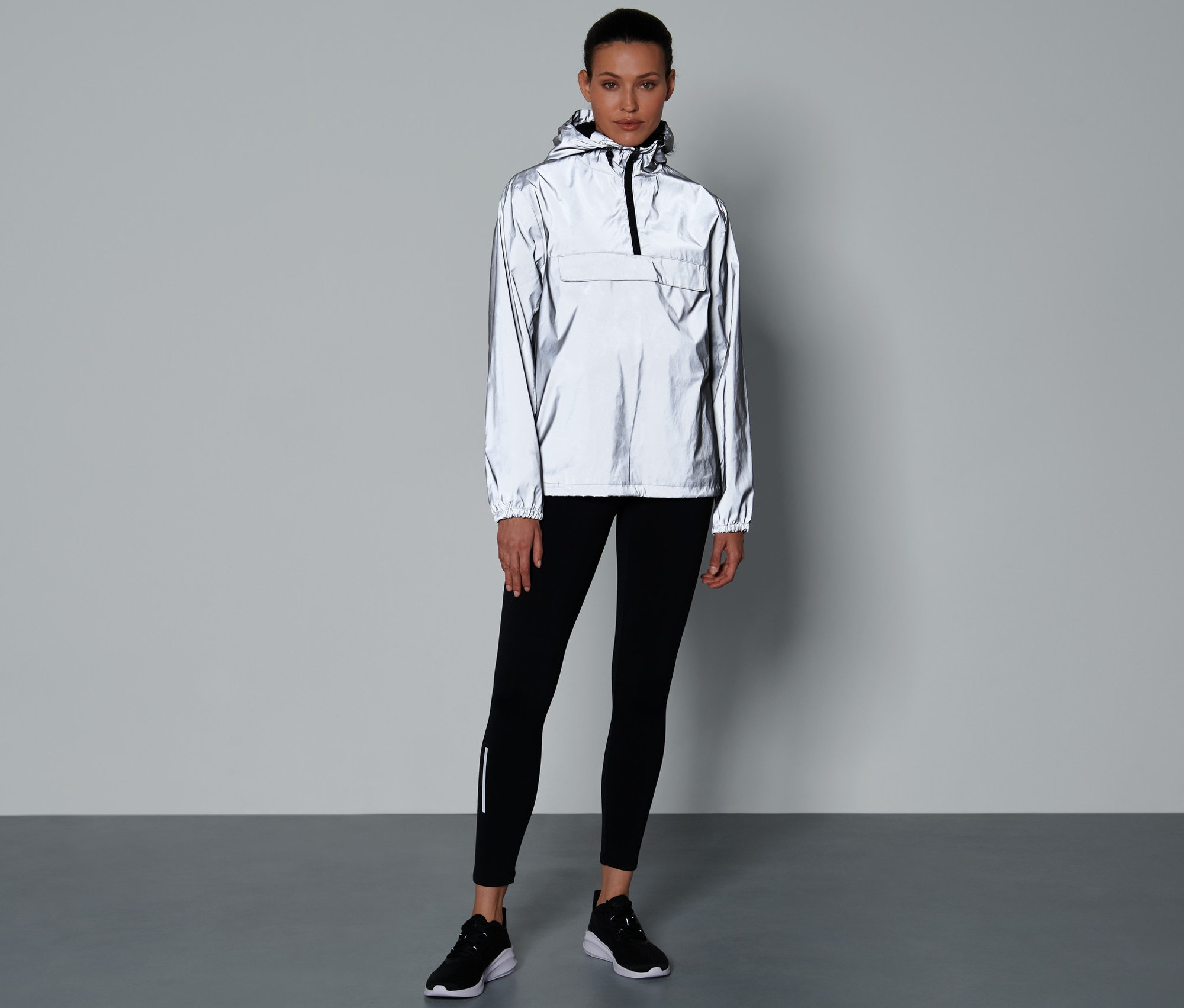 Une femme debout portant une veste coupe-vent réfléchissante, un legging noir et des chaussures.