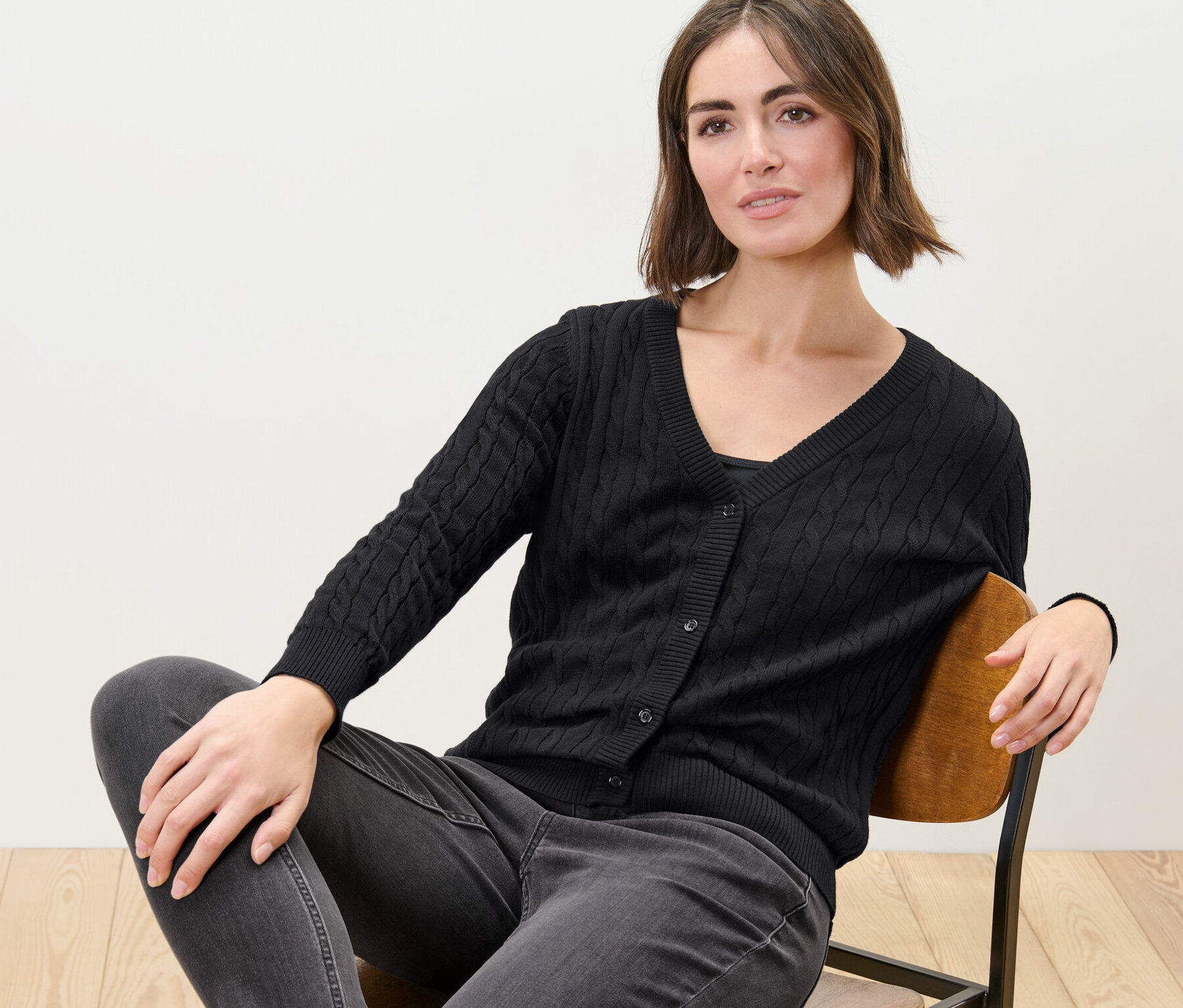 Une femme est assise sur une chaise et porte un cardigan noir en maille fine à motif torsadé.