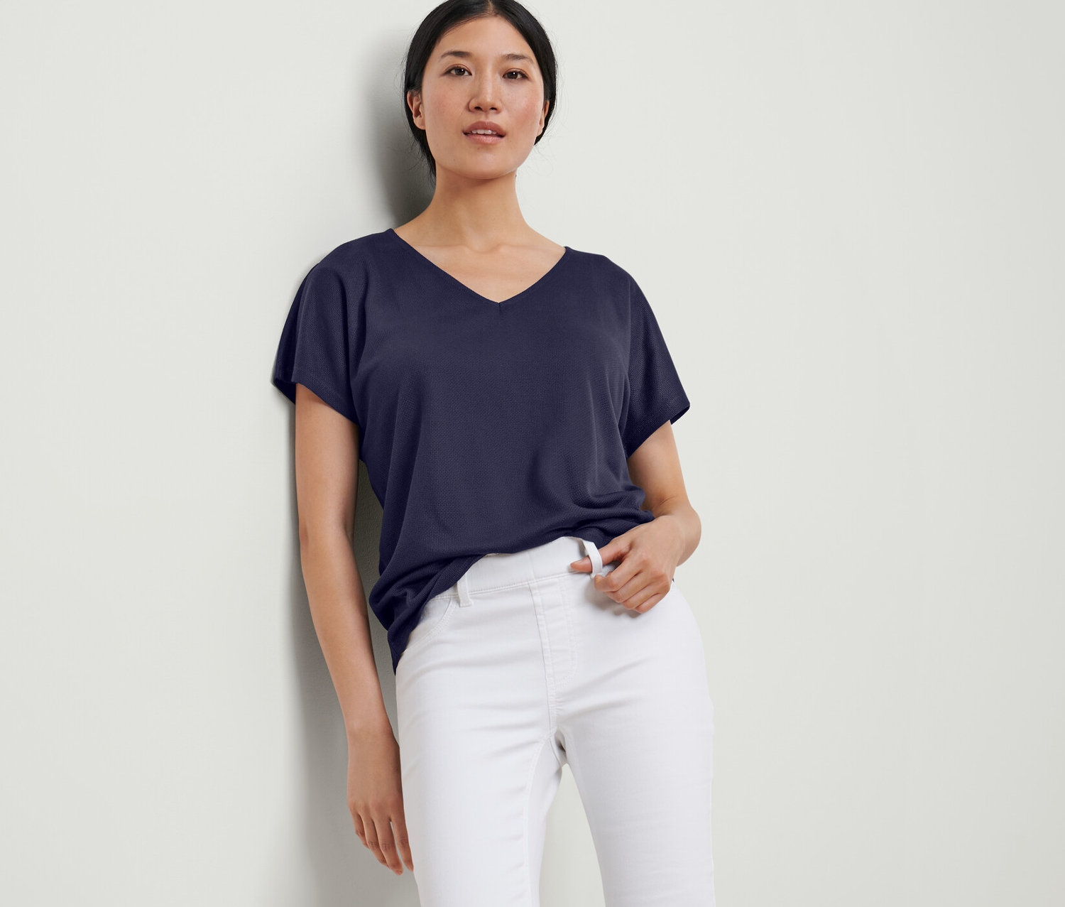 Une femme appuyée contre un mur porte une blouse structurée bleu foncé et un pantalon blanc.