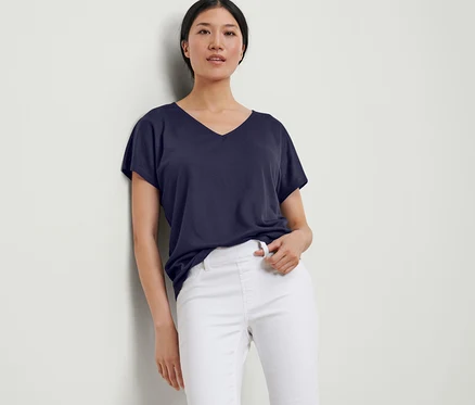 Une femme appuyée contre un mur porte une blouse structurée bleu foncé et un pantalon blanc.