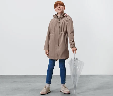 Une fille debout tenant un parapluie transparent. Elle porte un trench-coat tout-temps pour enfant, un jean et des baskets.