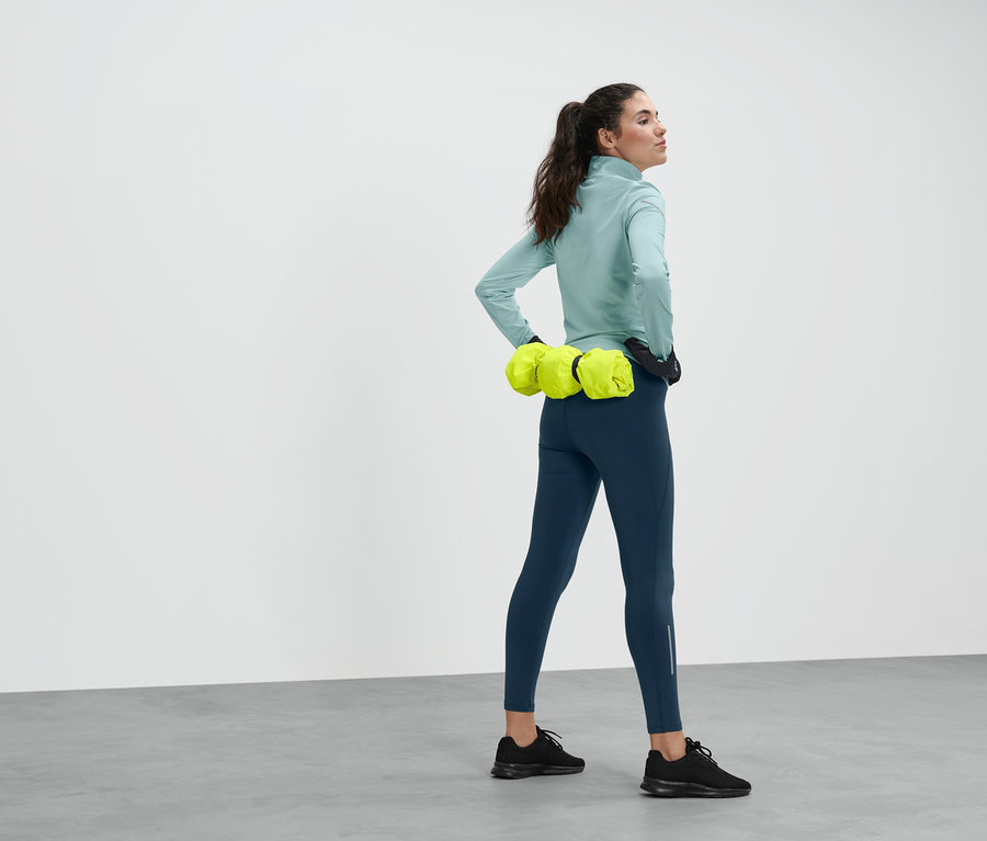 Femme posant en legging bleu, haut turquoise et chaussures de course noires. Autour de la taille, elle porte une veste de sport jaune.