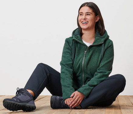 Une femme est assise sur un plancher en bois et porte une jaquette en maille polaire à capuche vert chiné et un pantalon thermique.