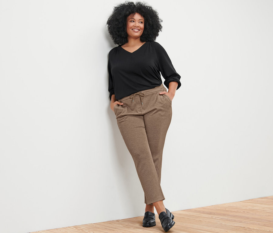 Femme appuyée contre un mur portant une blouse avec fronces et un pantalon de jogging beige.
