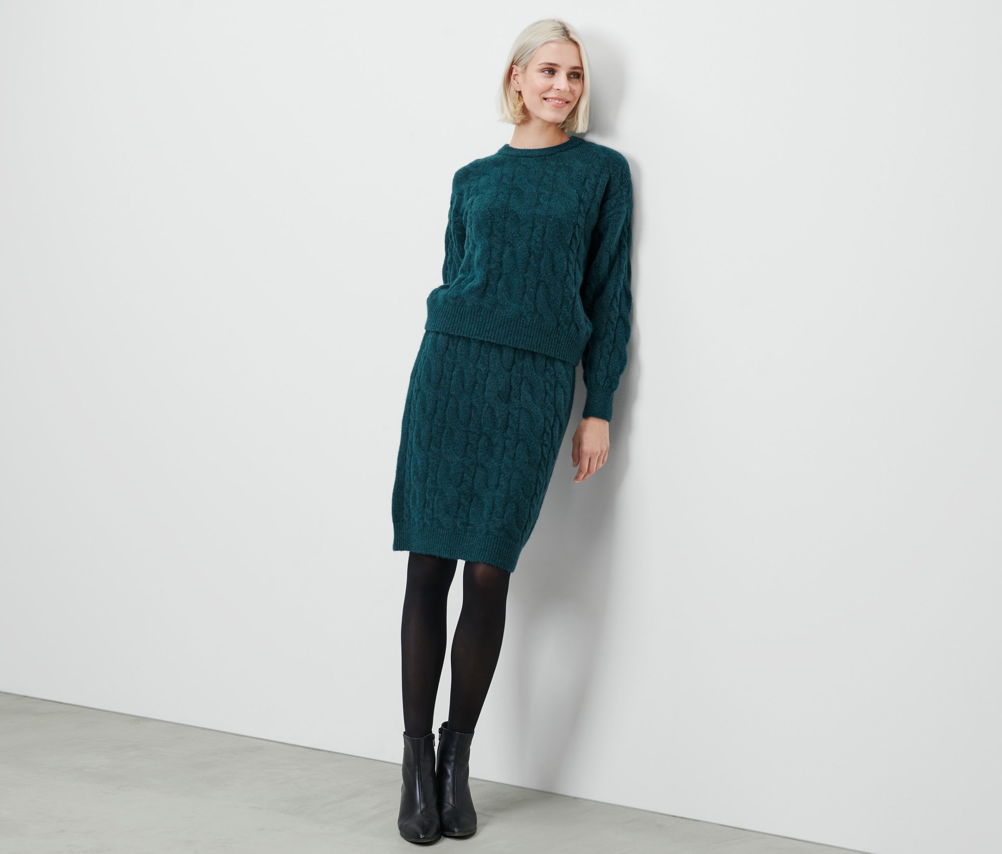 Une femme pose en jupe tricotée pétrole, pull, collant noir et bottes.