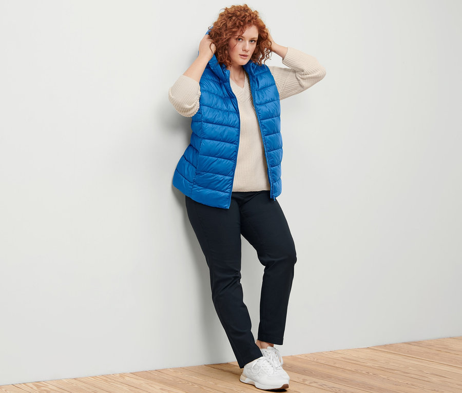Femme posant en gilet matelassé à capuche, pull en maille blanc cassé et pantalon en bengaline bleu foncé.