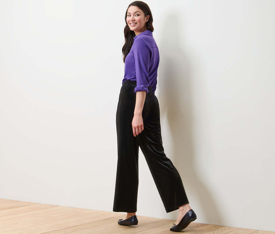 Une femme marche et regarde par-dessus son épaule, elle porte une chemise violette et un pantalon palazzo en velours noir.