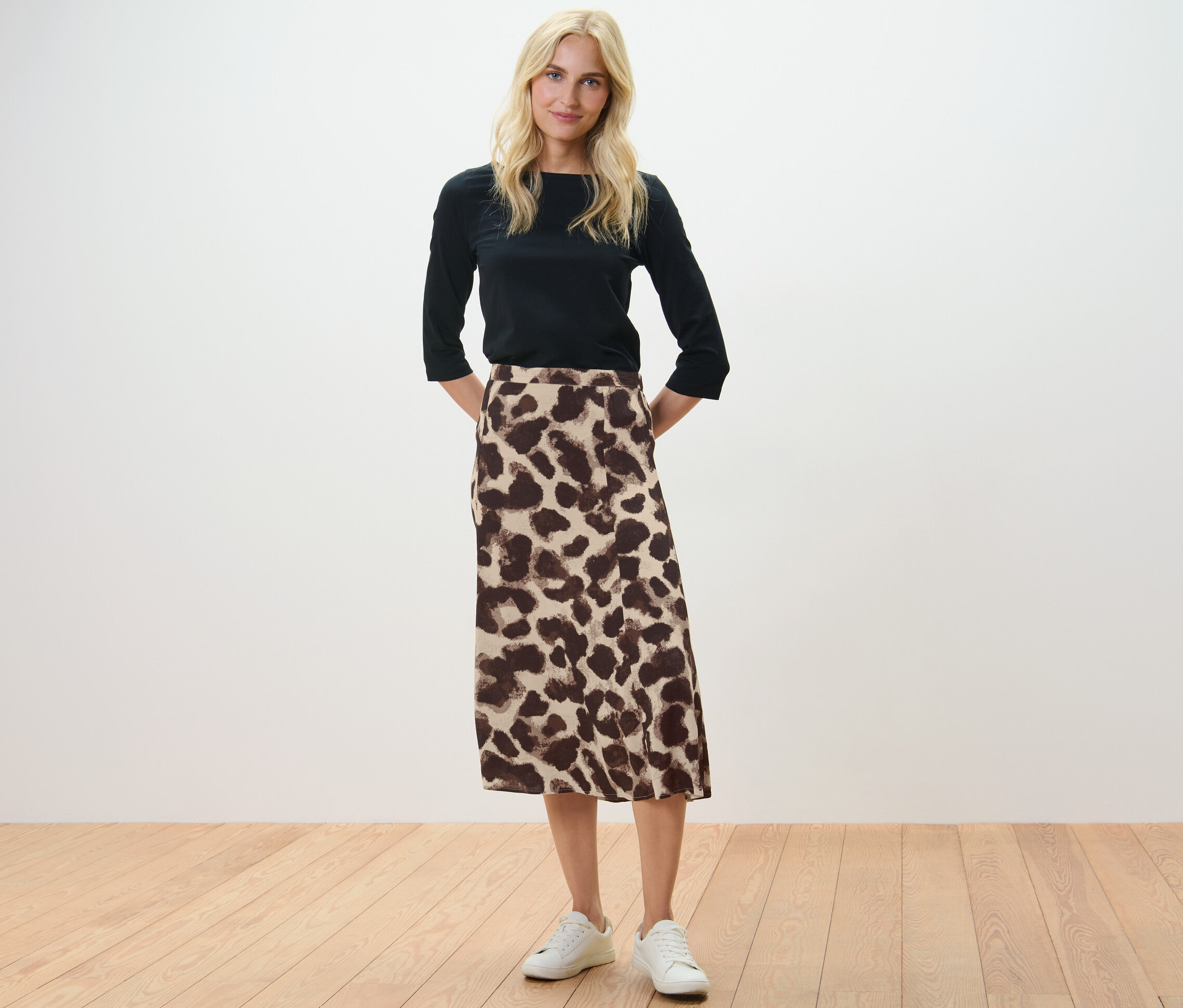 Une femme blonde se tient debout, les mains derrière le dos. Elle porte un chemisier noir et une jupe midi imprimée.