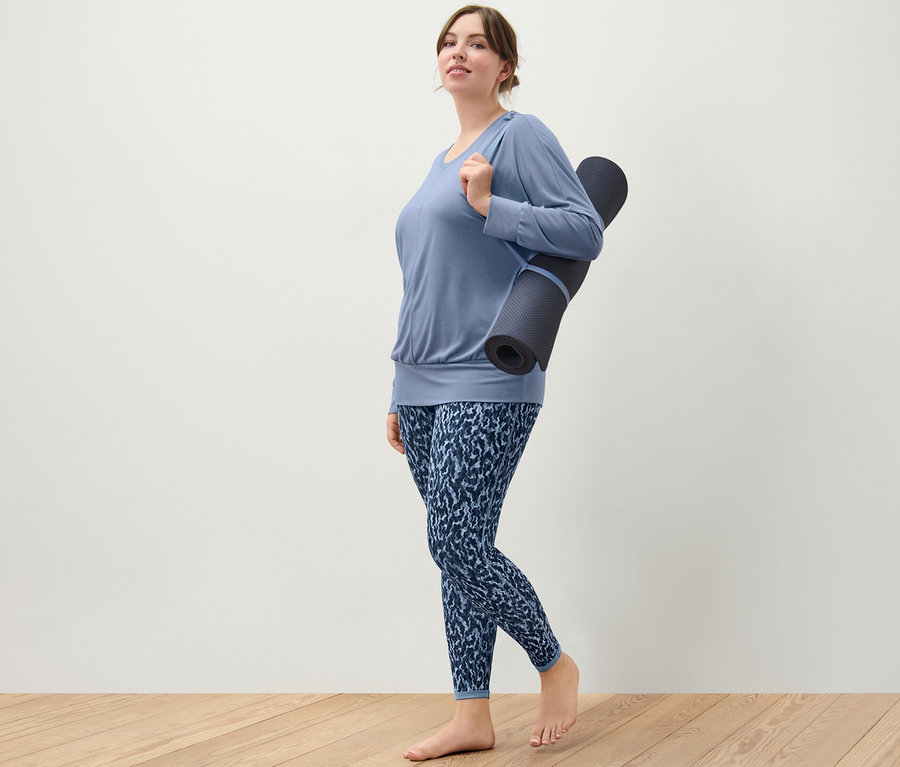 Femme portant un t-shirt de sport et de yoga bleu et un legging de sport réversible bleu/bleu clair, tenant un tapis de yoga sur l'épaule.