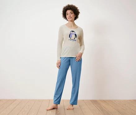 Une femme debout porte un haut de pyjama beige avec un motif de pingouin et un pantalon de pyjama bleu.