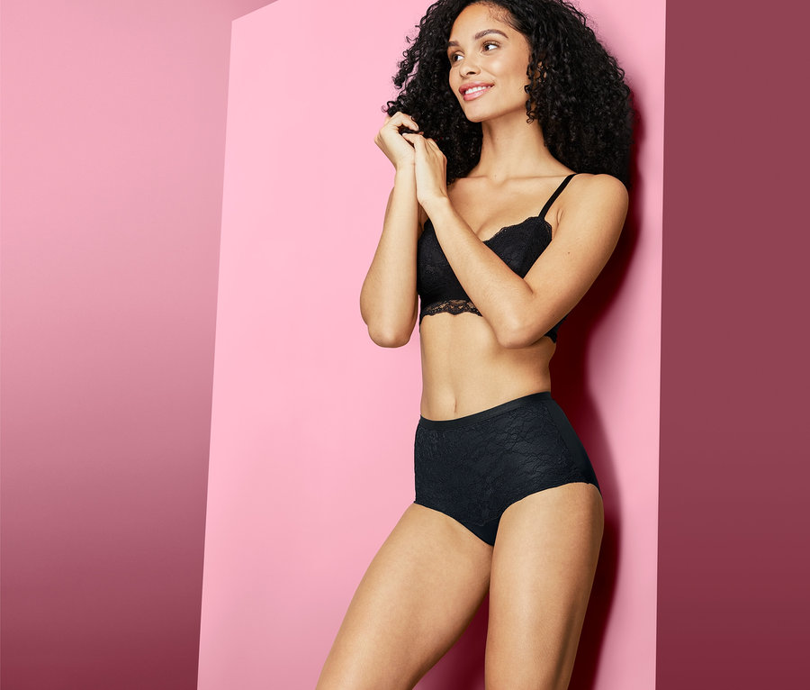 Femme aux cheveux bouclés foncés posant en culotte modelante noire et soutien-gorge en dentelle devant un fond rose.