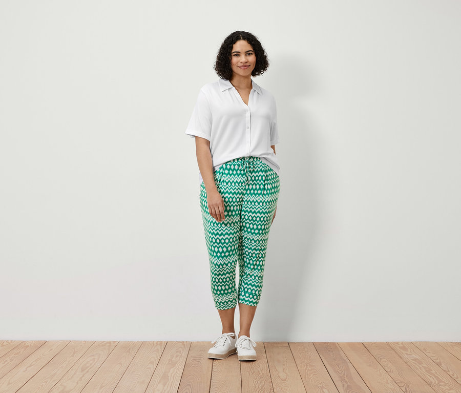 Une femme debout porte un pantalon 3/4 en jersey imprimé et une blouse blanche avec patte de boutonnage.