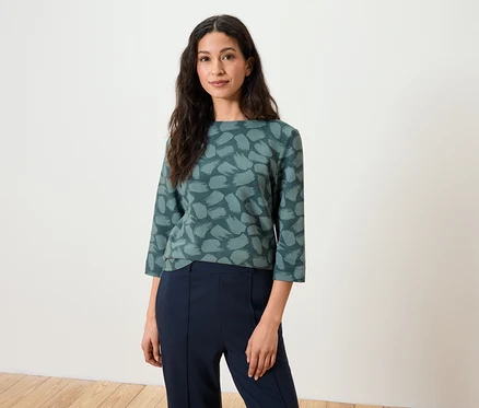 Femme aux cheveux bruns posant dans un haut à manches trois-quarts et un pantalon bleu marine.
