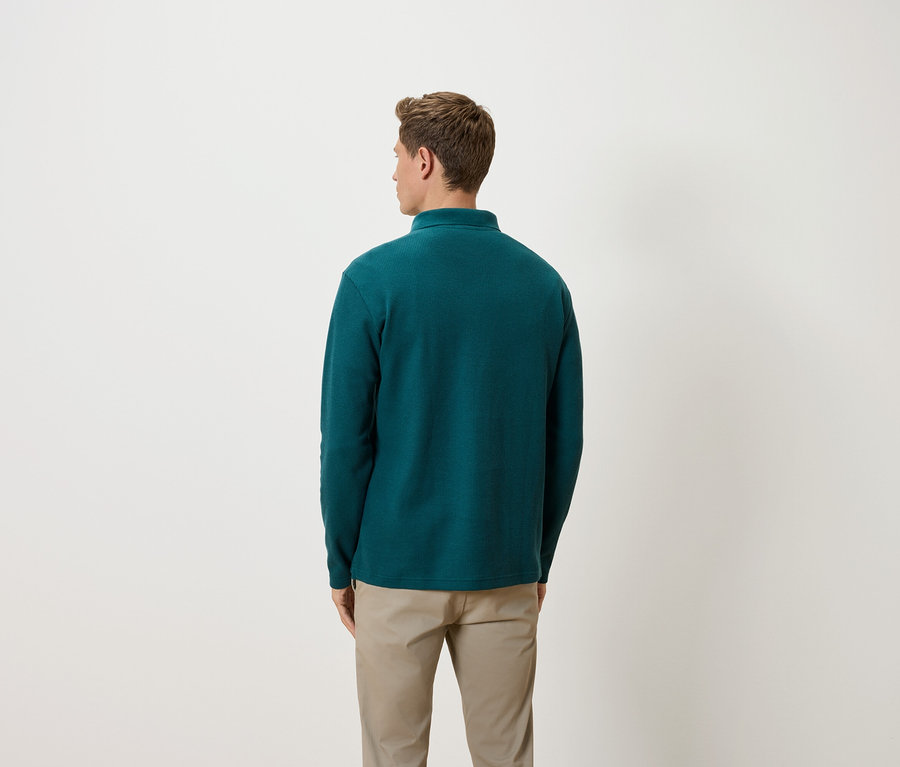 Un homme vu de dos portant un pull vert et un pantalon beige.