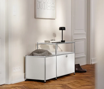 Dans un couloir, un meuble TV bas en métal blanc »CN3« avec un compartiment à rabat et un plateau en verre, sur lequel reposent un livre et une lampe.