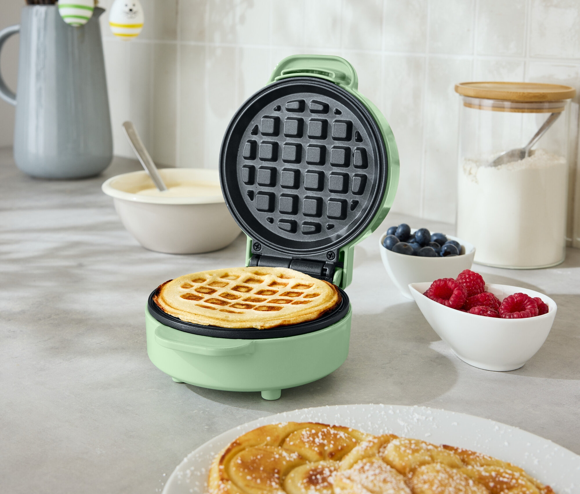 Un gaufrier ouvert, rond et vert menthe avec une gaufre à l'intérieur se trouve sur un comptoir de cuisine. À côté, il y a deux pâtisseries en spirale dans une assiette, un bol de myrtilles et un bol de framboises.