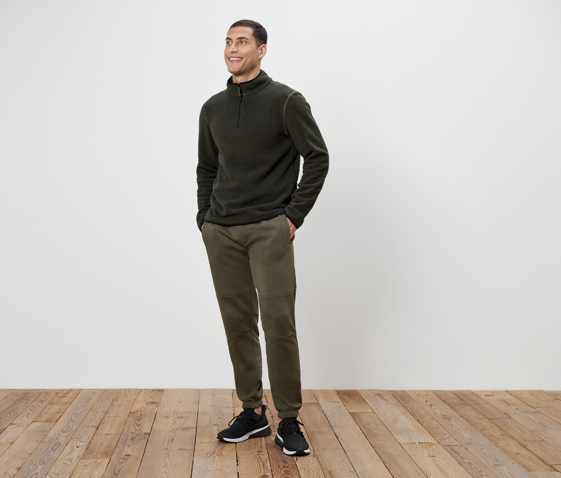 Un homme portant un pantalon vert olive et un pull en polaire gris foncé à fermeture éclair, debout sur un plancher en bois.