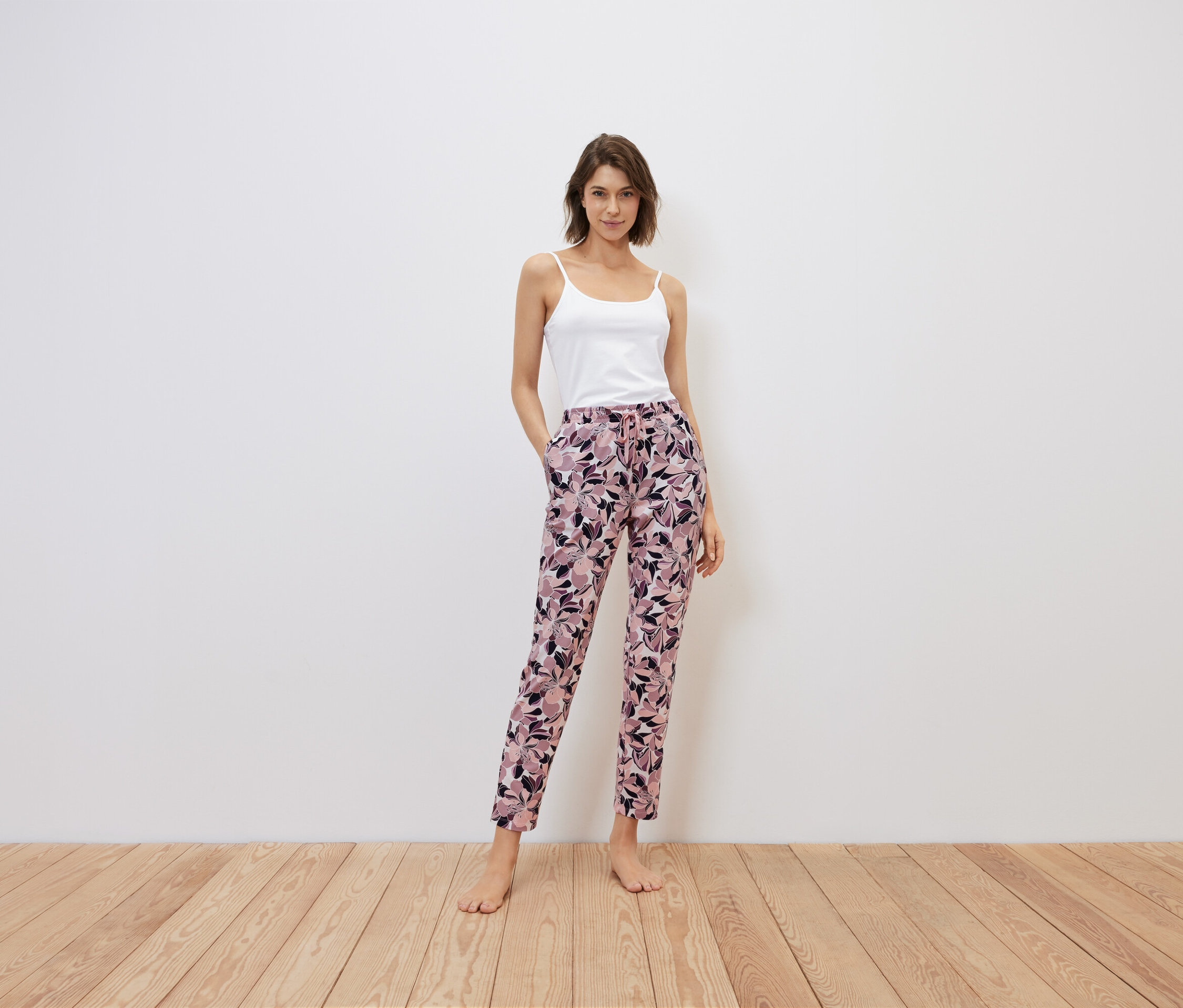 Une femme se tient debout, les mains dans les poches d'un pantalon à fleurs, haut blanc.