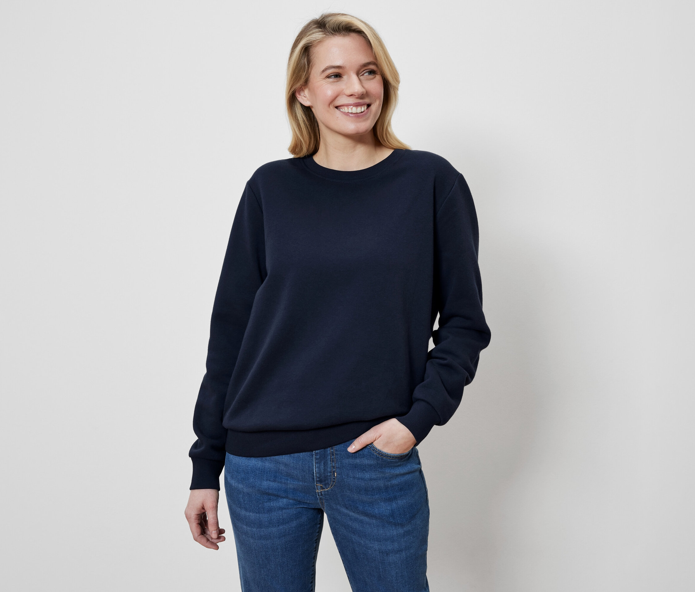 Une femme blonde, la main dans la poche de son jean, sourit et porte un sweat-shirt bleu marine.