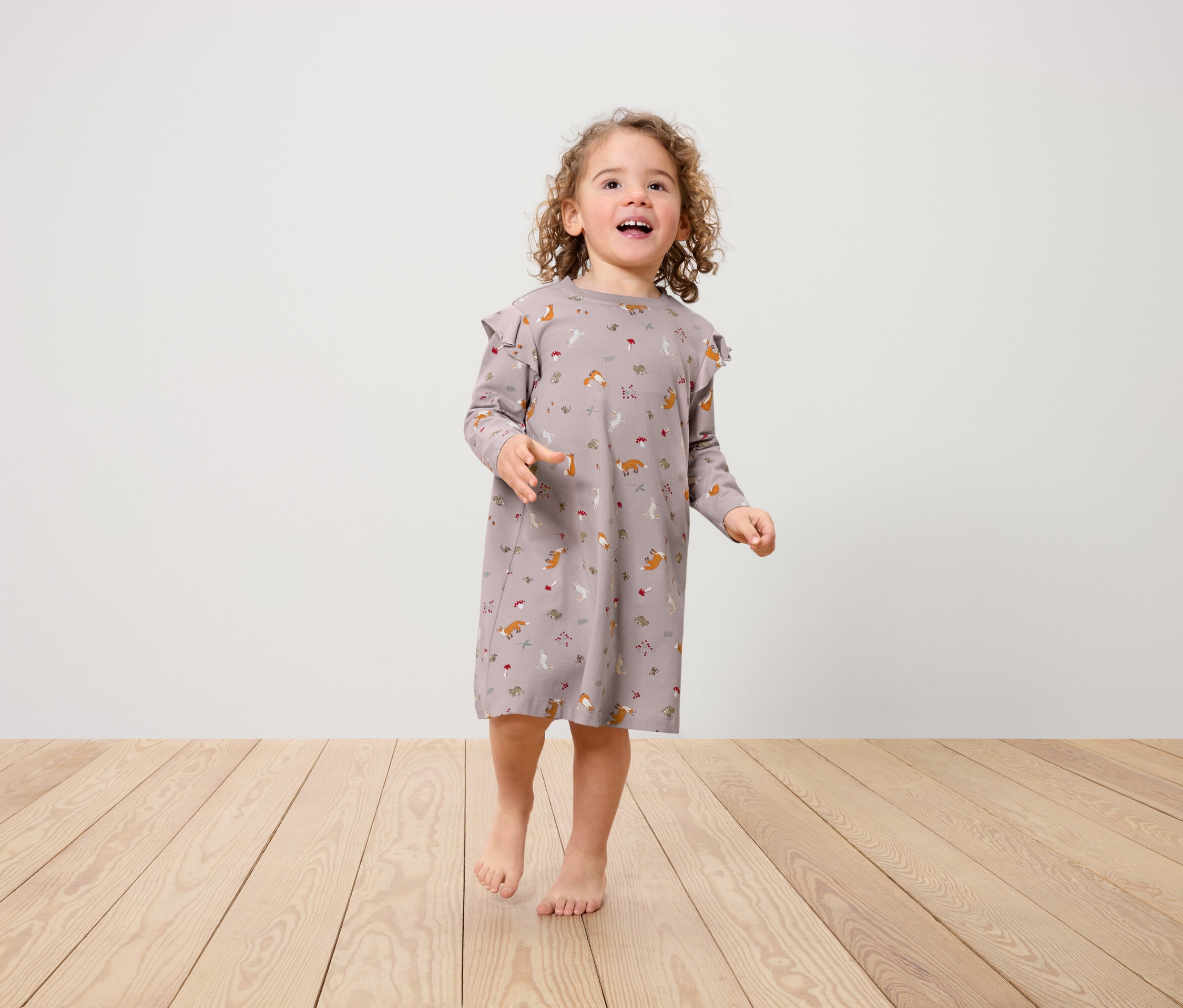 Une petite fille portant une robe à motifs de renards avec des volants aux épaules se tient sur un plancher en bois.