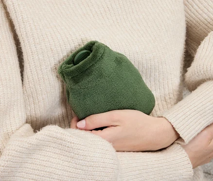 Une femme en pull tient une bouillotte verte et douce dans ses mains.