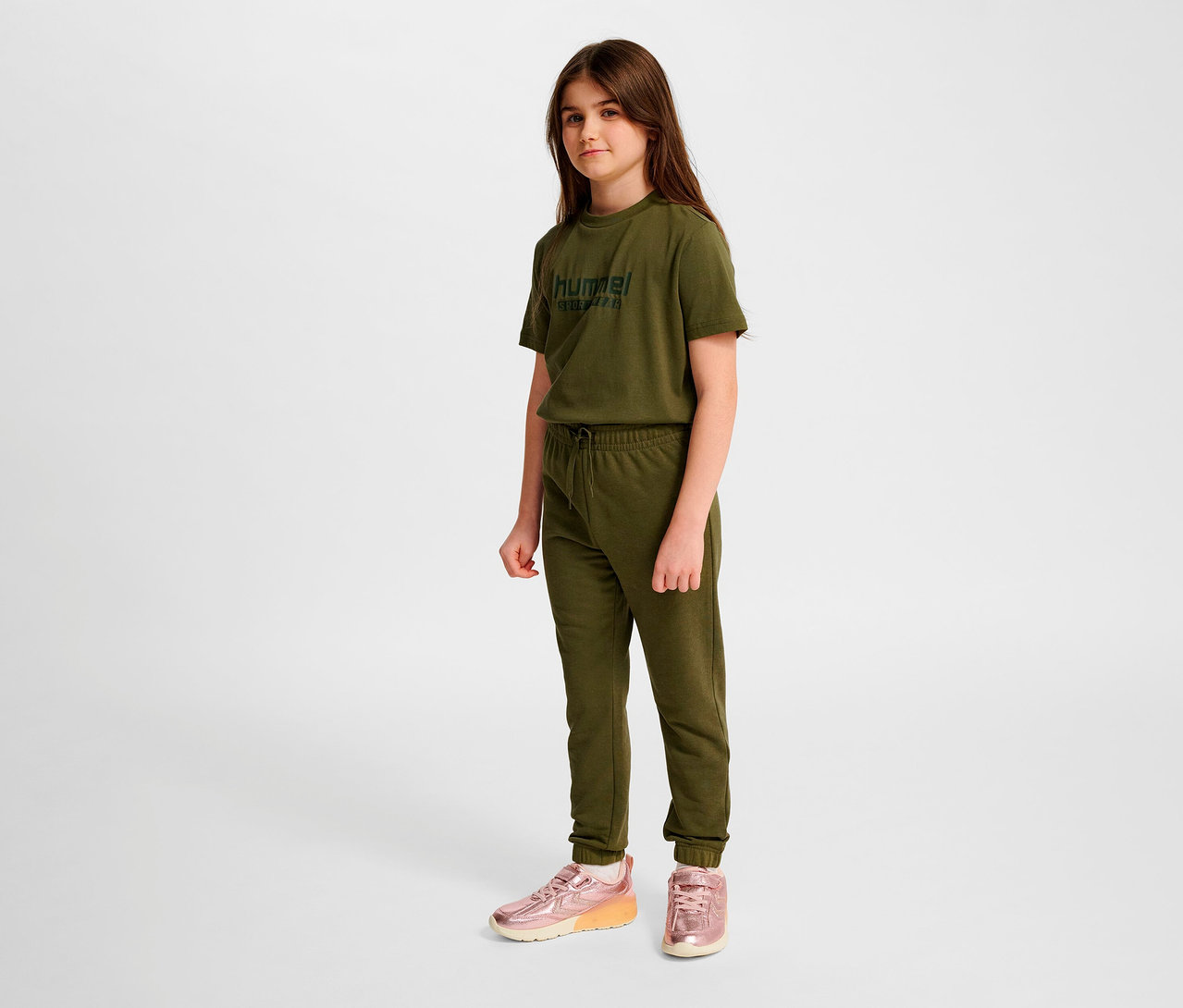Une fille debout portant un t-shirt vert, un pantalon régulier HUMMEL HMLJR Base vert et des chaussures roses.