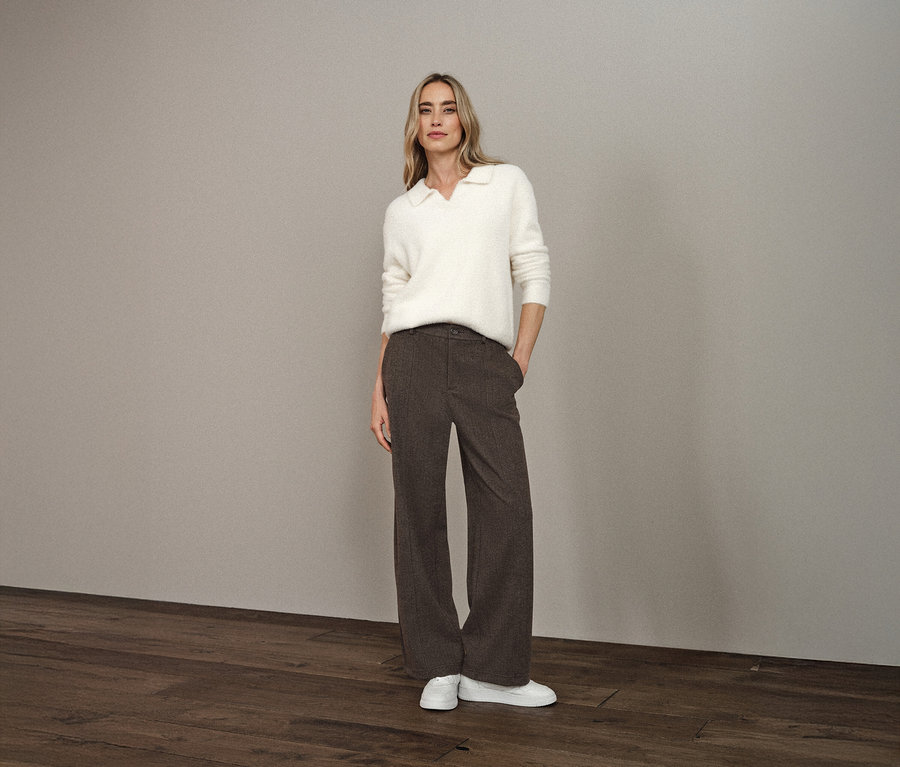 Une femme se tient debout, les mains dans les poches de son pantalon, et porte un pull en maille à col polo Street One, un pantalon marron et des chaussures blanches.