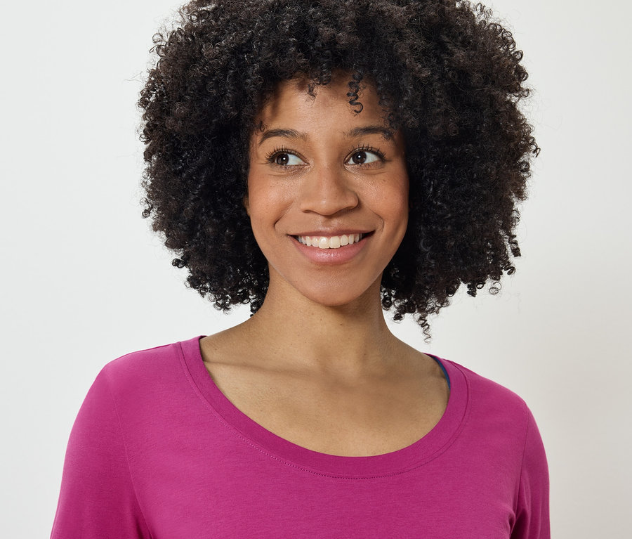 Portrait d'une femme souriante avec une coiffure afro portant un t-shirt rose, regardant vers la gauche.