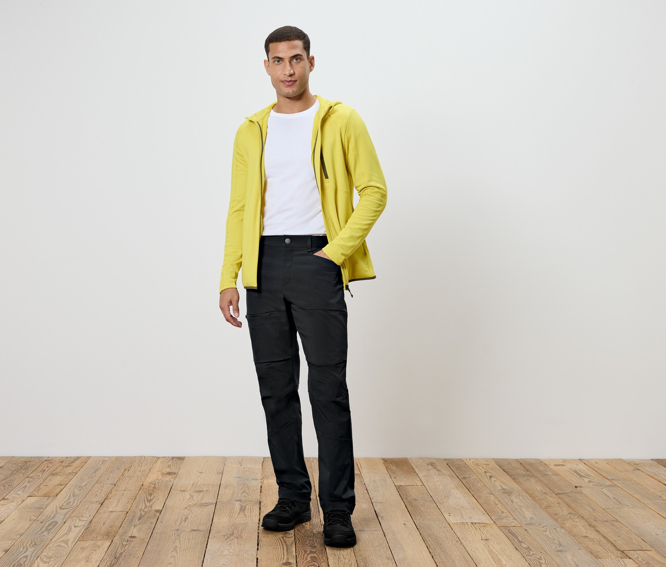 Un homme debout sur un plancher en bois. Il porte un pantalon noir, un t-shirt blanc et un sweat à capuche jaune.