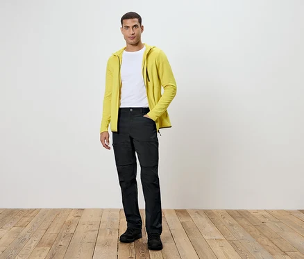 Un homme debout sur un plancher en bois. Il porte un pantalon noir, un t-shirt blanc et un sweat à capuche jaune.