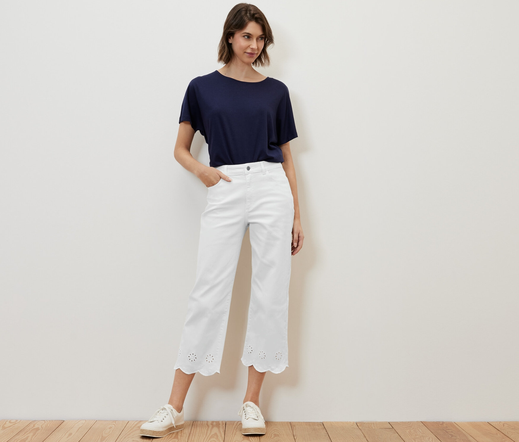 Une femme pose en jean blanc avec broderie ajourée et un t-shirt bleu.