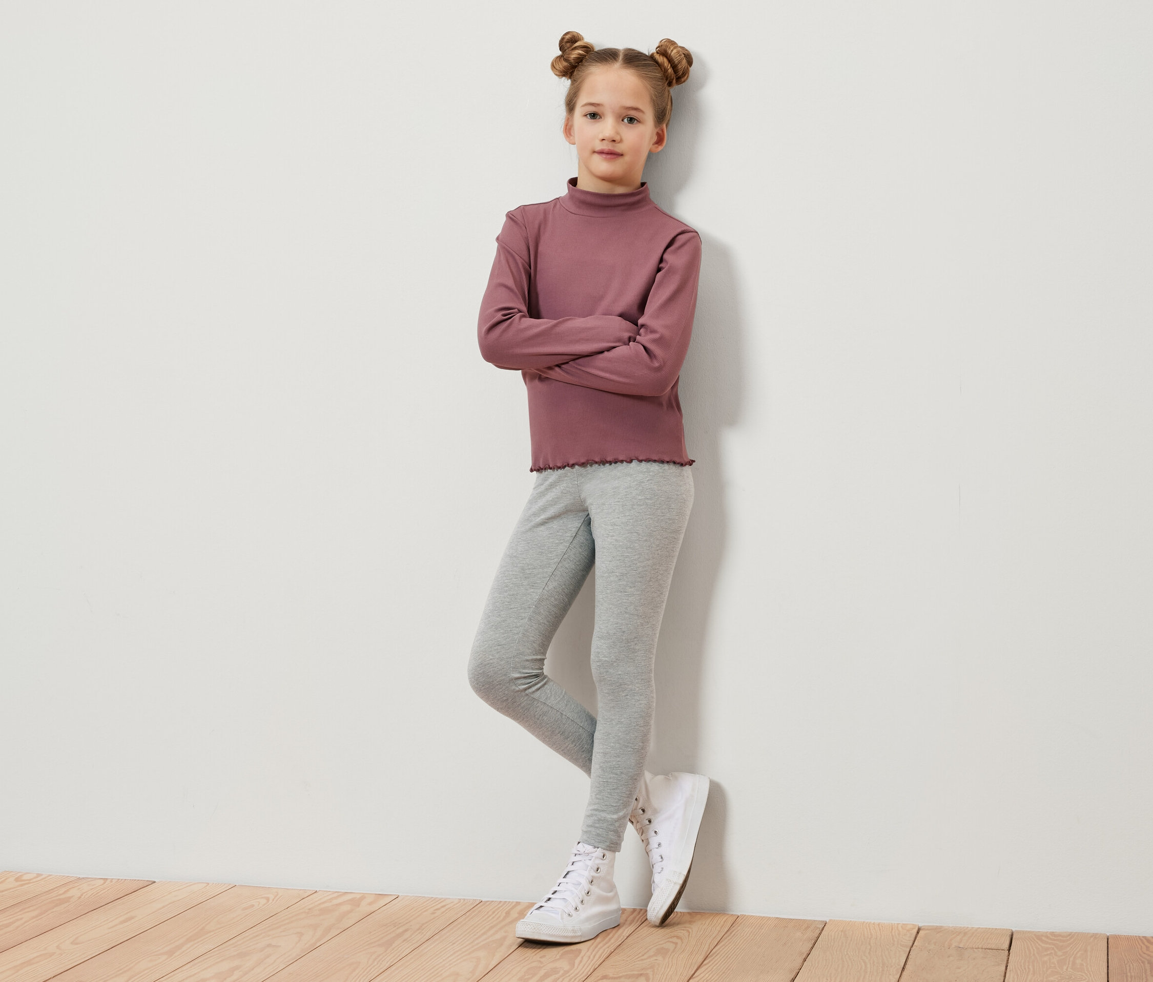 Une jeune fille aux cheveux bruns coiffés en chignons croisés se tient debout, les bras croisés. Elle porte un t-shirt à manches longues bordeaux, un legging gris et des baskets blanches.