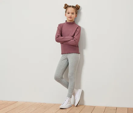 Une jeune fille aux cheveux bruns coiffés en chignons croisés se tient debout, les bras croisés. Elle porte un t-shirt à manches longues bordeaux, un legging gris et des baskets blanches.