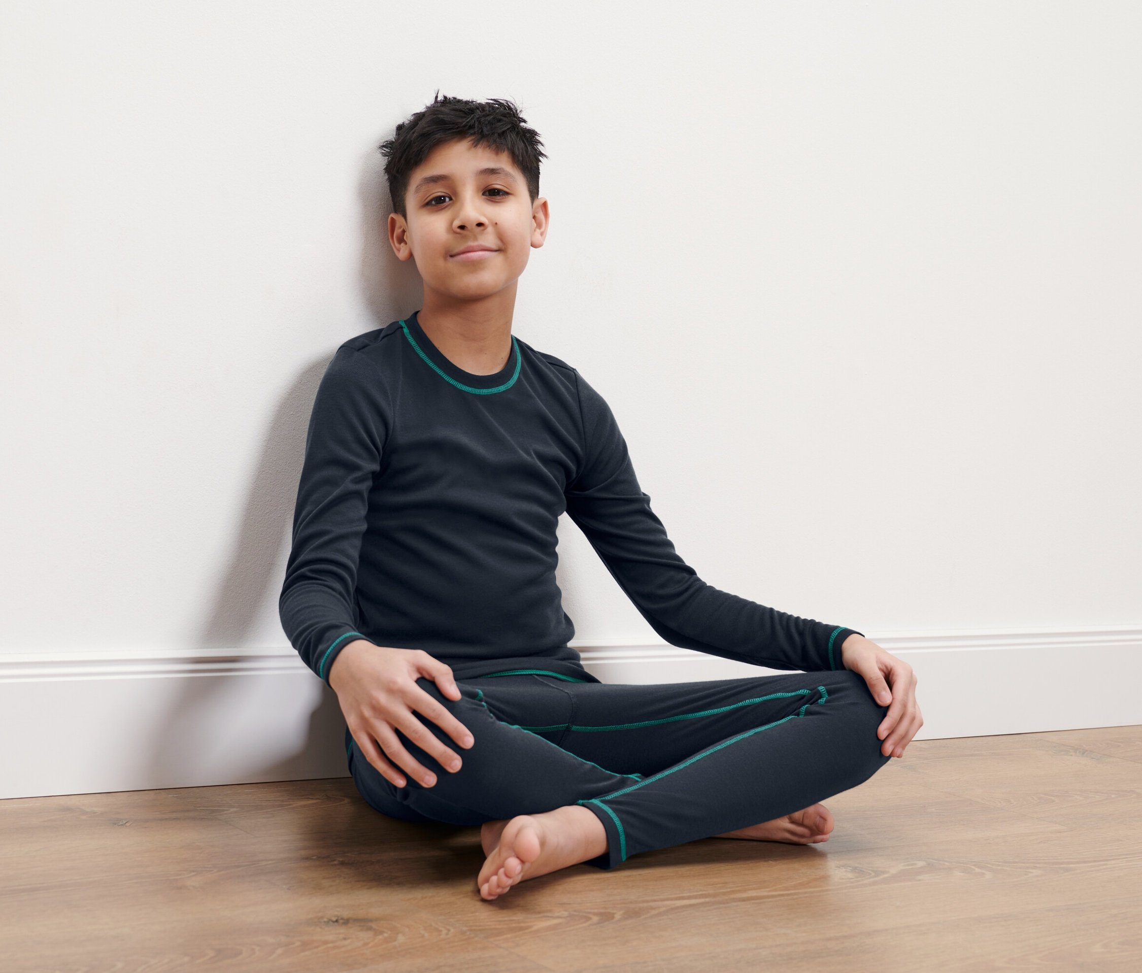Un garçon est assis les jambes croisées sur le sol, adossé à un mur blanc. Il porte des sous-vêtements thermiques bleu foncé.