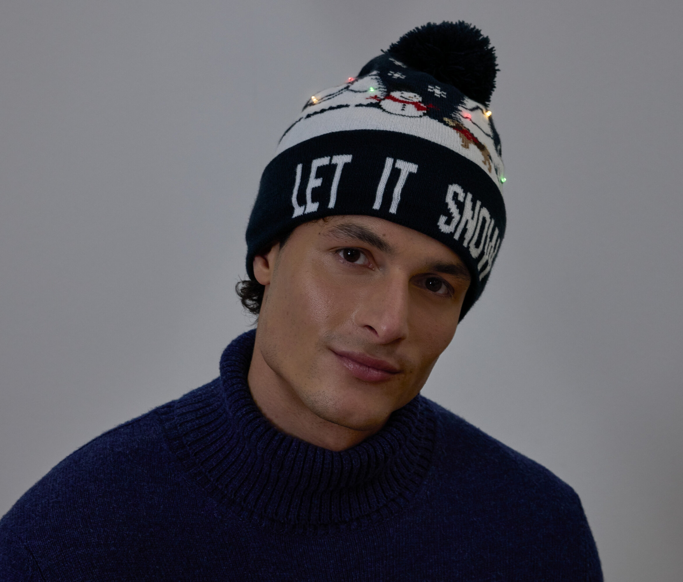 Portrait d'un homme portant un bonnet de Noël avec l'inscription LET IT SNOW et de petites lumières.